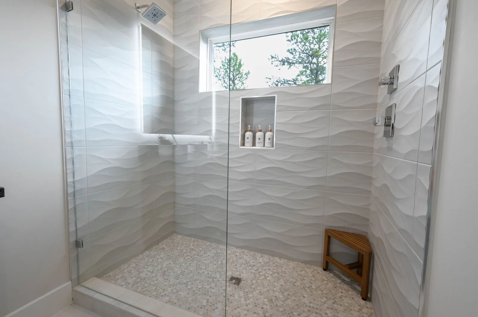 Walk-in shower at The Aroha with pebble floor, wave-textured tile walls, and wooden bench