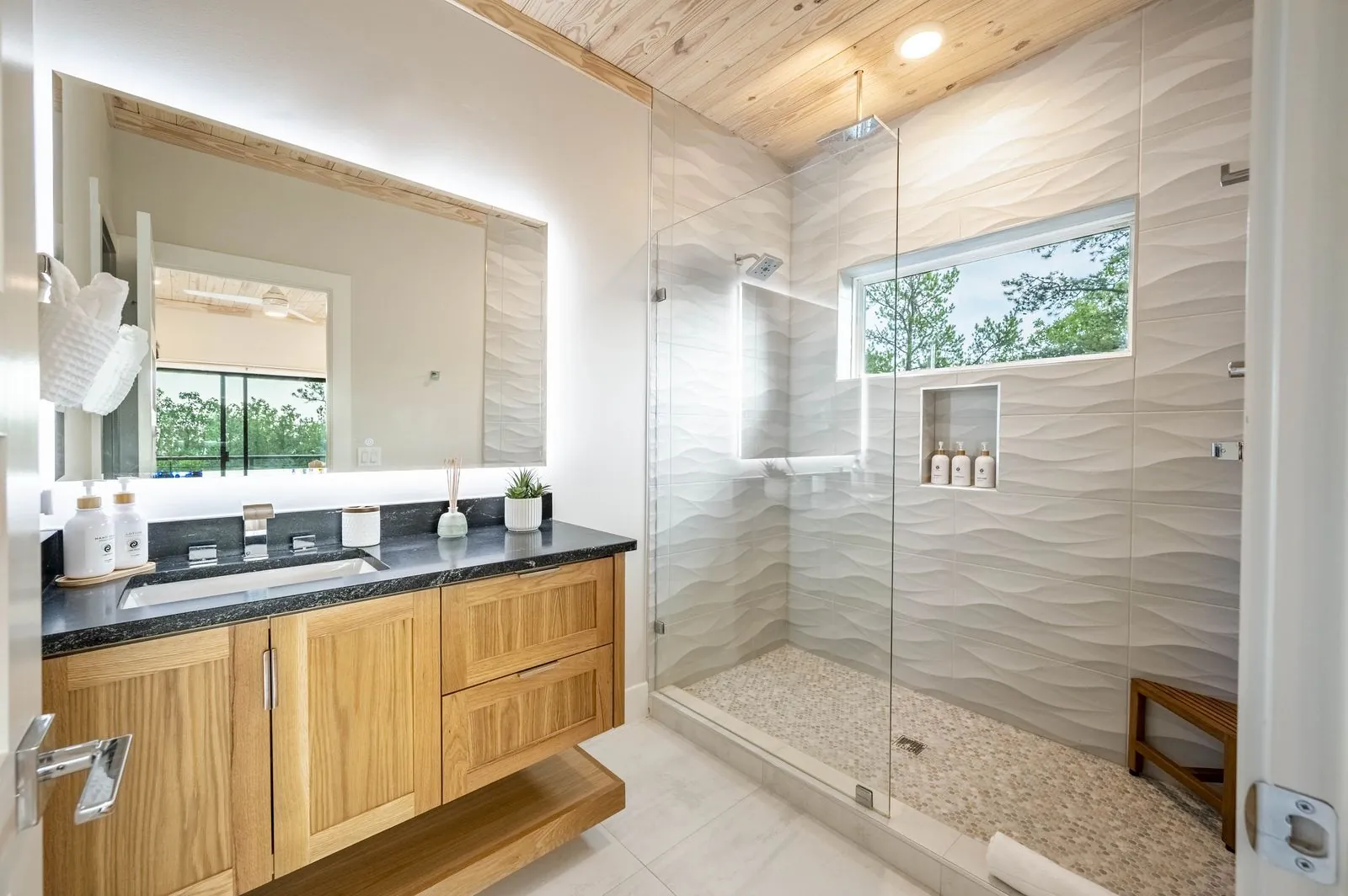Glass-enclosed walk-in shower at The Aroha with wave tile, pebble floor, and wooden bench