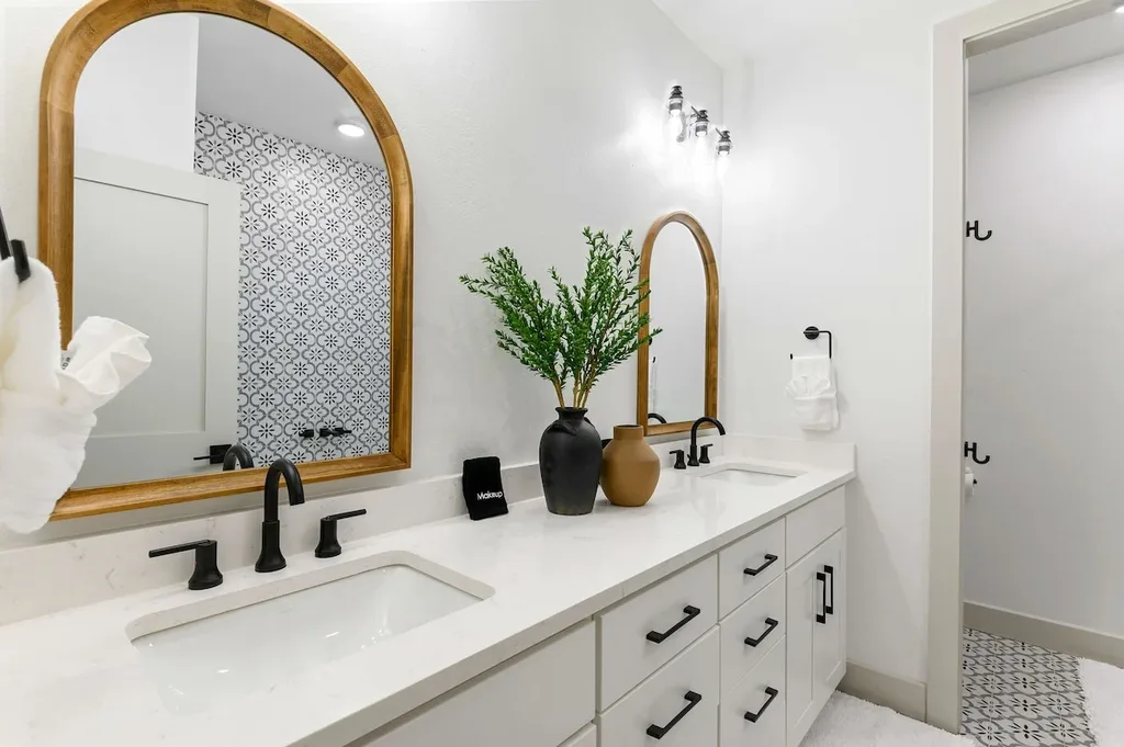 This private ensuite bathroom has a double vanity with white quartz countertop, black fixtures, and arched wood-framed mirrors, plus a separate shower area through the doorway.