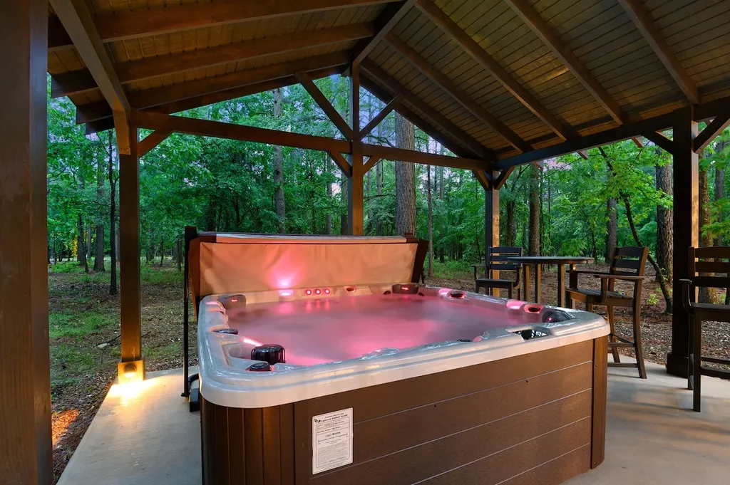 A private hot tub sits under a covered cabana with seating nearby, surrounded by trees at the back of the cabin.