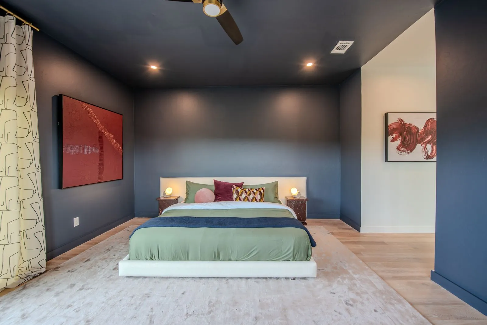 Blue-walled bedroom at The Jade showing window with natural light, ceiling fan, and modern abstract artwork
