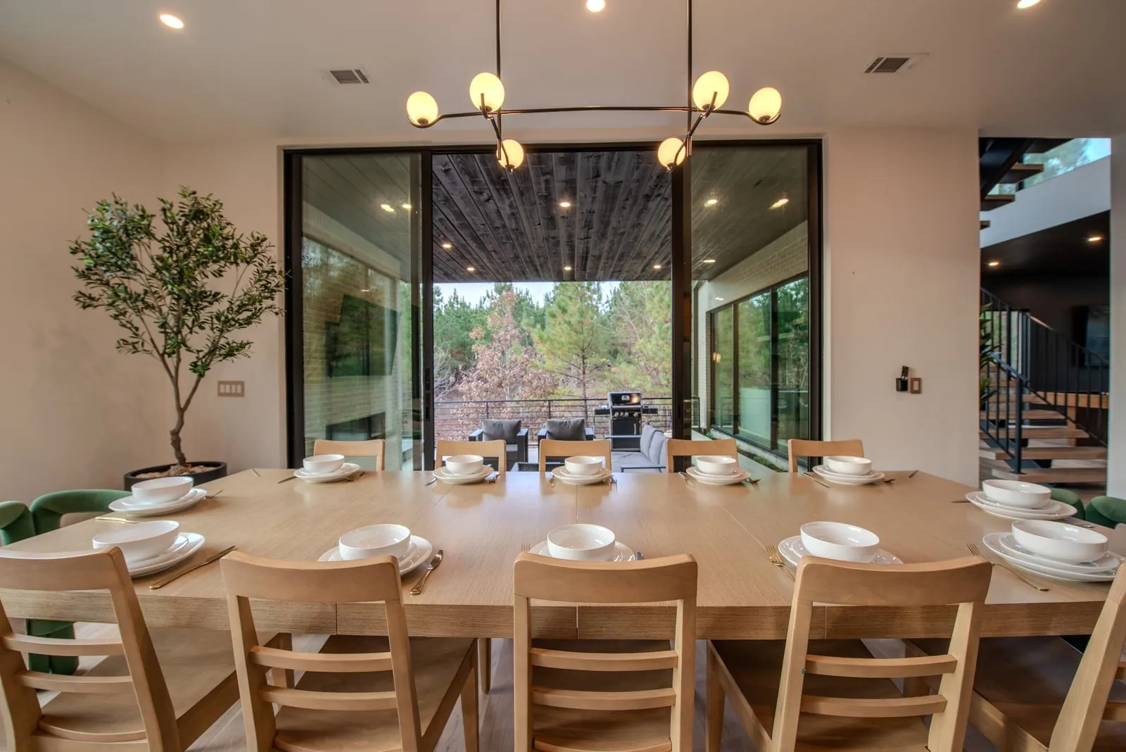 Dining table set with wood chairs beneath a brass chandelier with floor-to-ceiling windows overlooking forest at The Jade