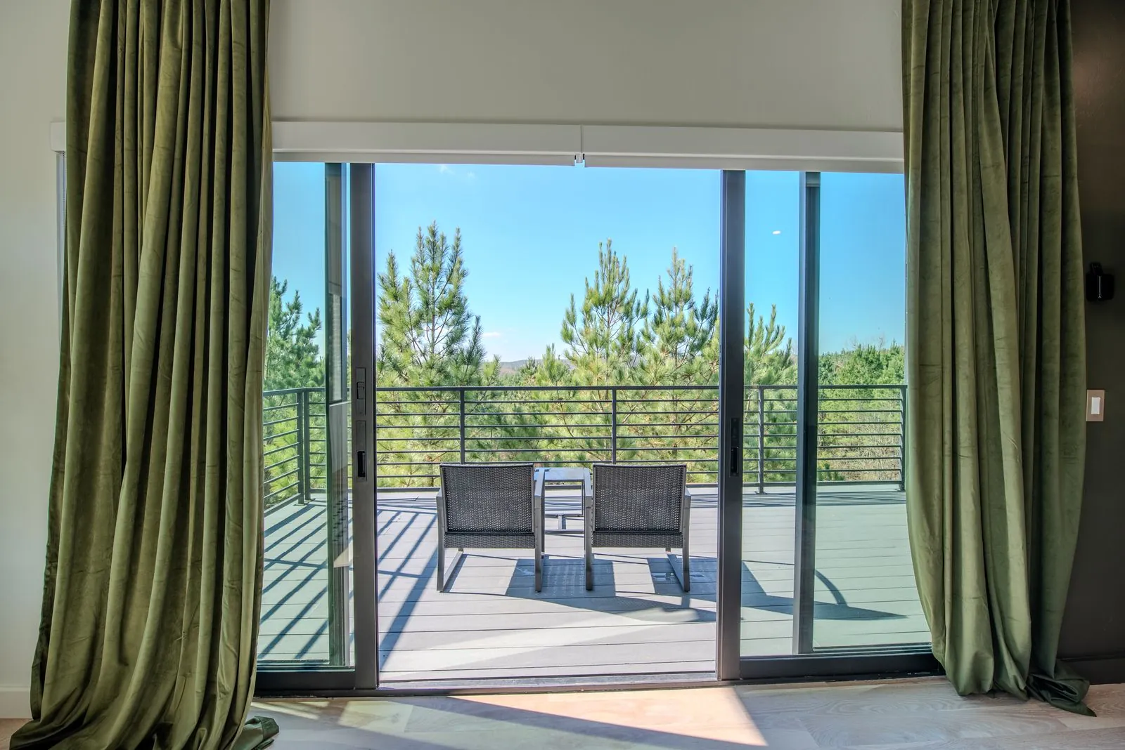 Primary bedroom sitting area at The Jade with green floor-length curtains, wall-mounted TV, and balcony access with forest views