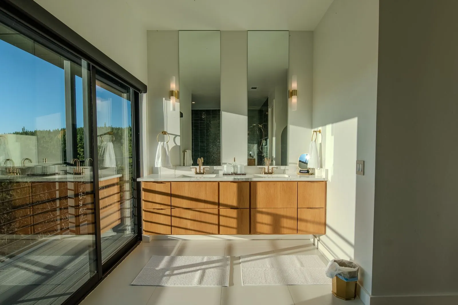 Double vanity bathroom at The Jade with large mirrors, warm wood cabinetry, modern sconces, and forest view through windows