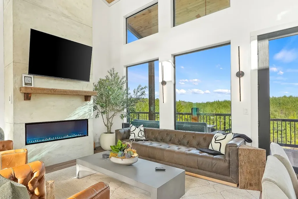 Sunlit living room with sectional, linear fireplace, television, and floor-to-ceiling forest windows